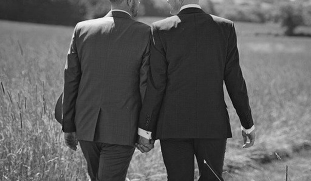 a black and white image showing a couple of man in suites, holding hands, out in an open field, represents a politician in a sex scandal